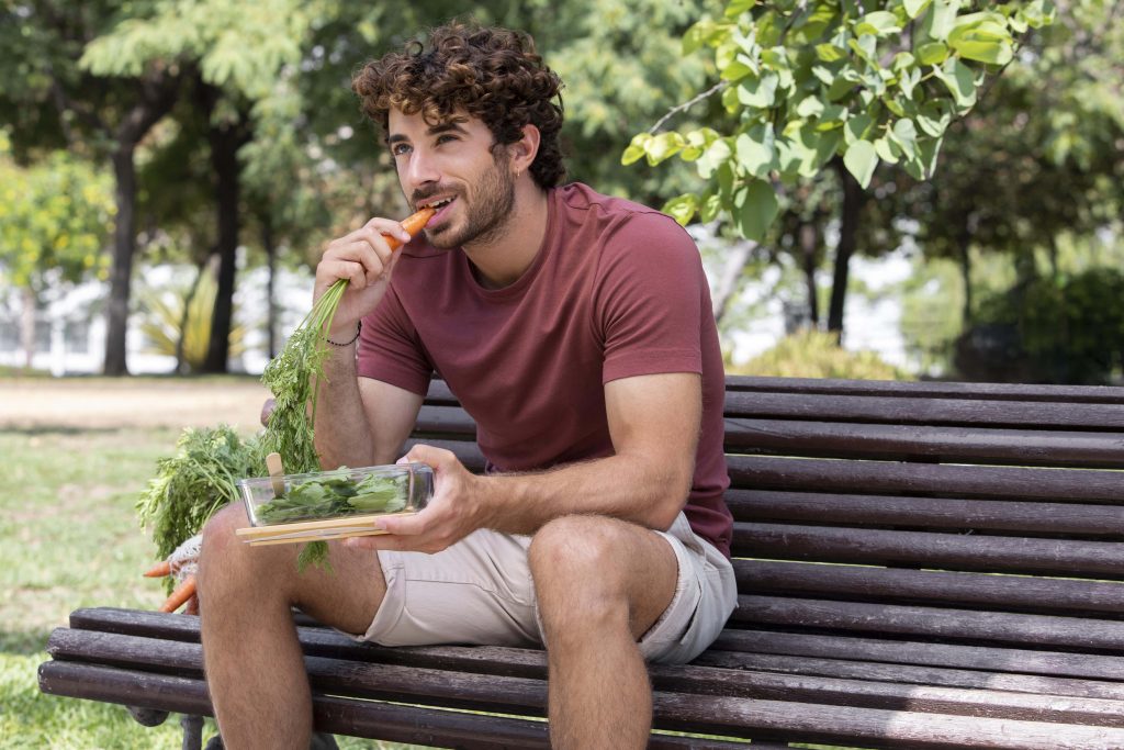 A man bites a carrot while sitting on a bench in a park.