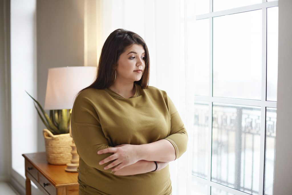 obese woman standing worried looking out the glass window thinking about how to lose weight