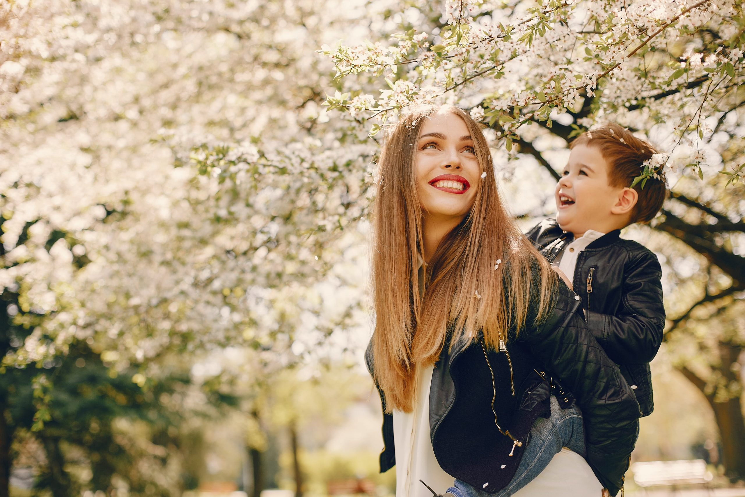 woman carries her little kid on her back and smiles looking at the blooming trees in summer