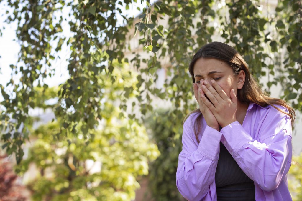 woman sneezing in spring as she enters a park with lots of trees and flowers