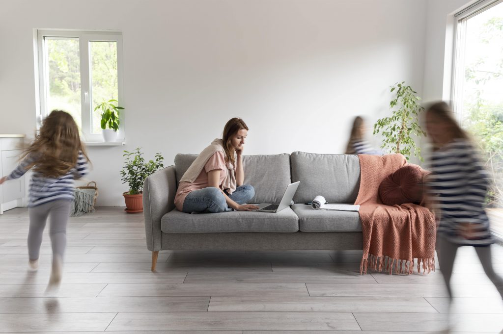 woman working seriously on lap top in living room sofa and her kids keep running around her and playing