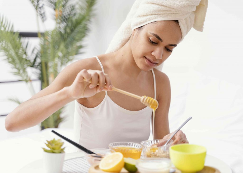 woman in bath robe making a paste with honey placed in bowl. nearby is a bowl of honey and some herbs 