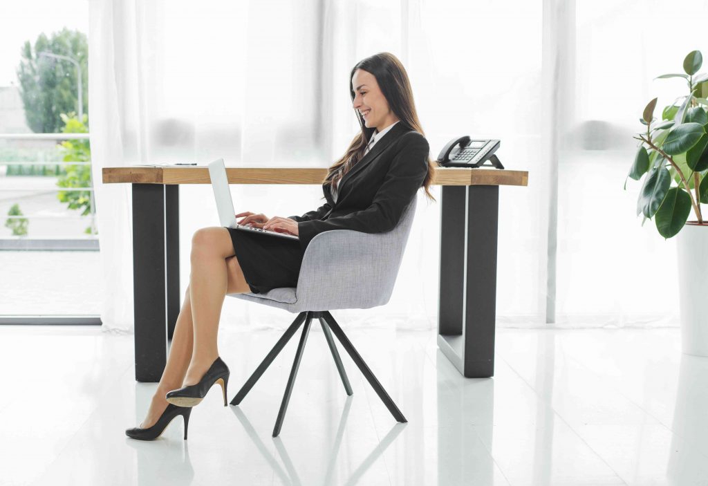 professional woman in office attire, sitting cross legged in chair holding a lap top in the thighs and working