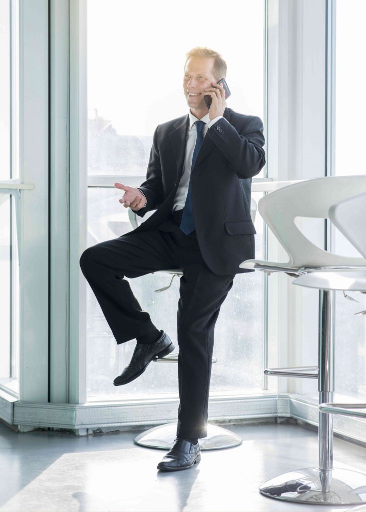 man in professional attire, sitting on high stool, standing on one leg and while resting other leg on the chair, talking on the phone to someone