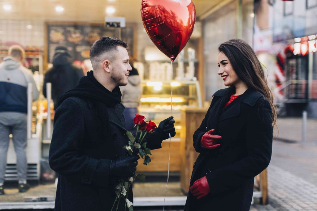 man showers woman with flowers and love balloon in public place, they both smile 