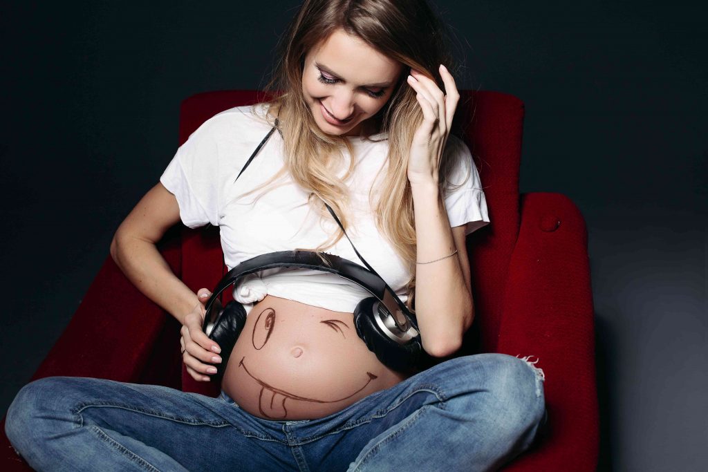 woman sitting on sofa with her pregnant exposed holding a headphone over the tummy